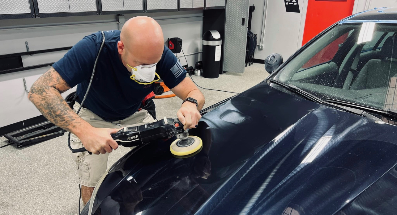 Close-up of a dual-action polisher refining dark paint under LED shop lighting